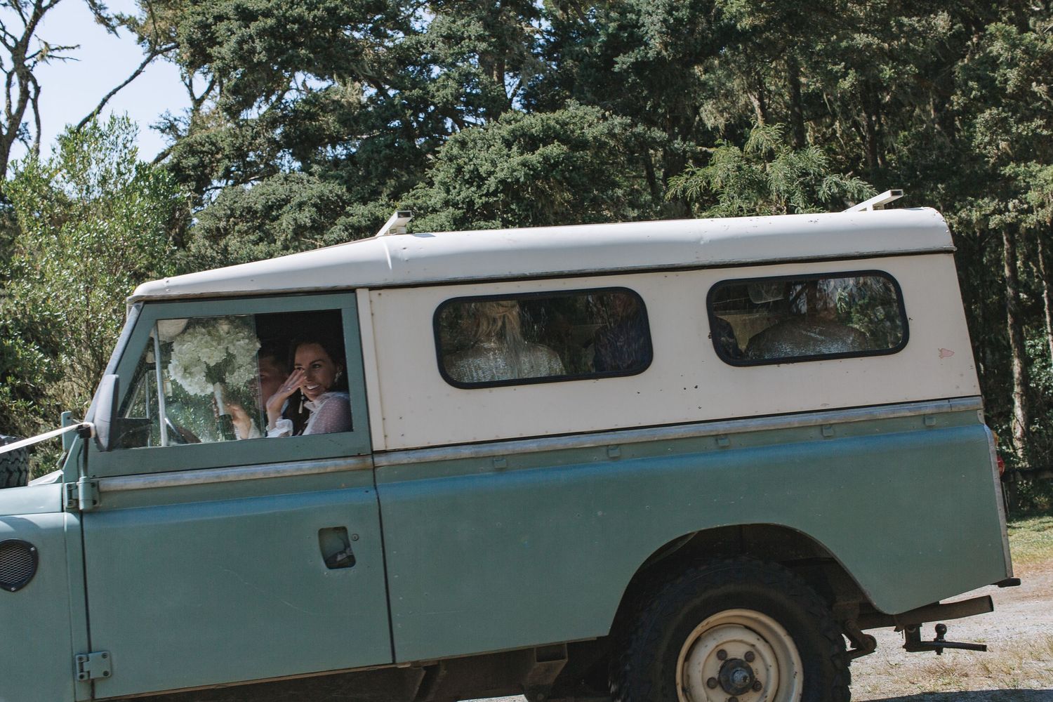 A vintage turquoise and white Land Rover Series truck parked under trees on a sunny day.