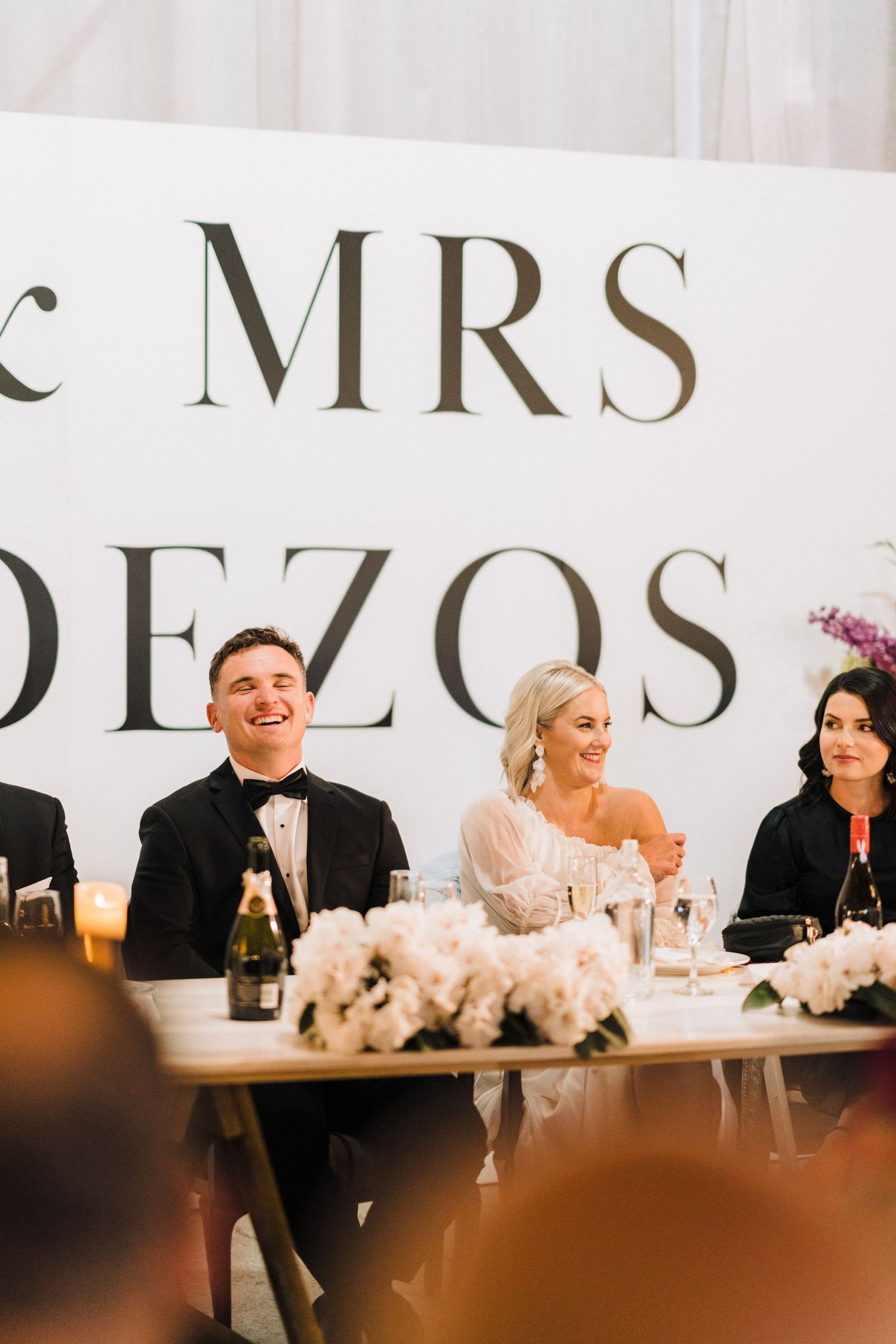 Wedding party sits at an elegant head table with floral centerpieces and illuminated 'MRS' signage in the background.