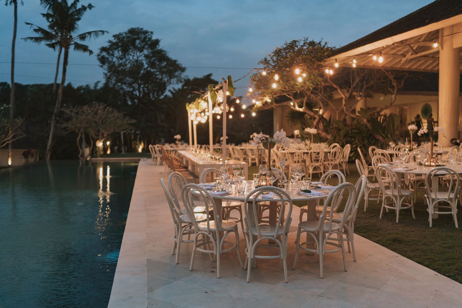 Elegant outdoor wedding reception setup with string lights and waterfront views at twilight.