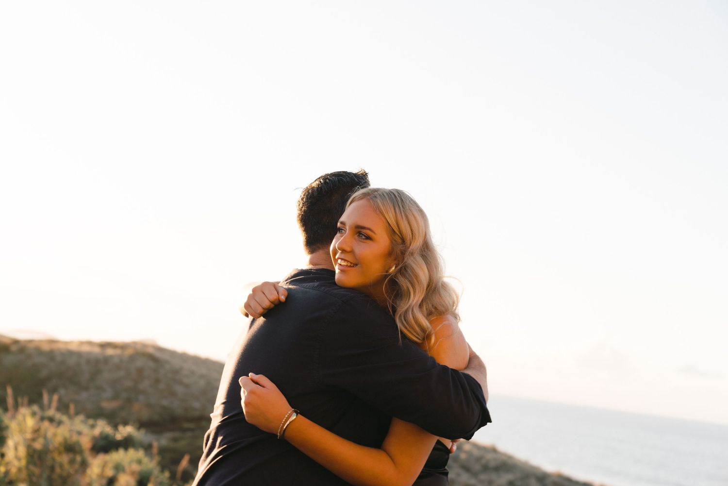 Two people embrace in a candid moment against a sunset backdrop on a coastal hillside.