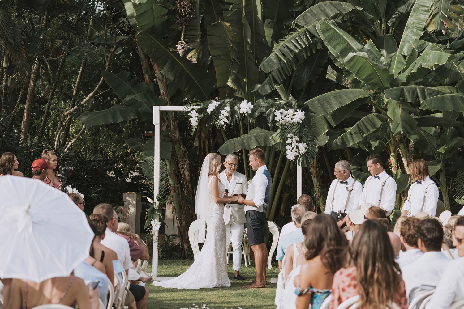 Wedding party members in white attire pose together in a lush tropical garden setting during a celebration.
