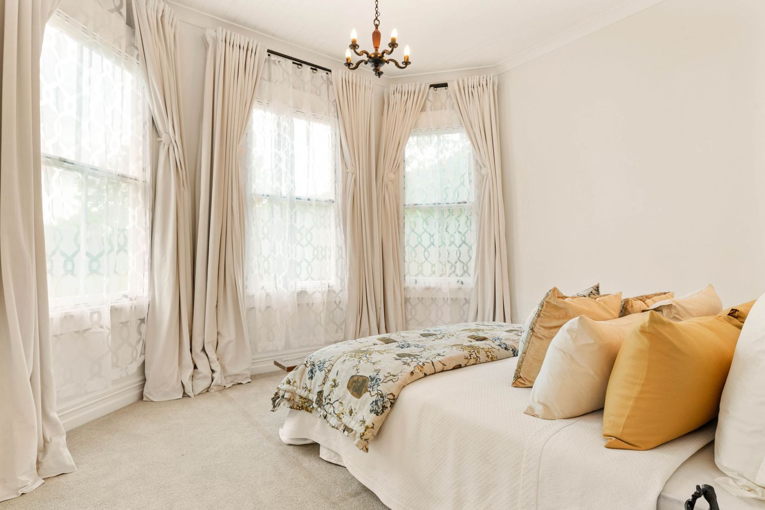 Bright bedroom with flowing cream curtains, bay windows, and white bedding with yellow accent pillows.