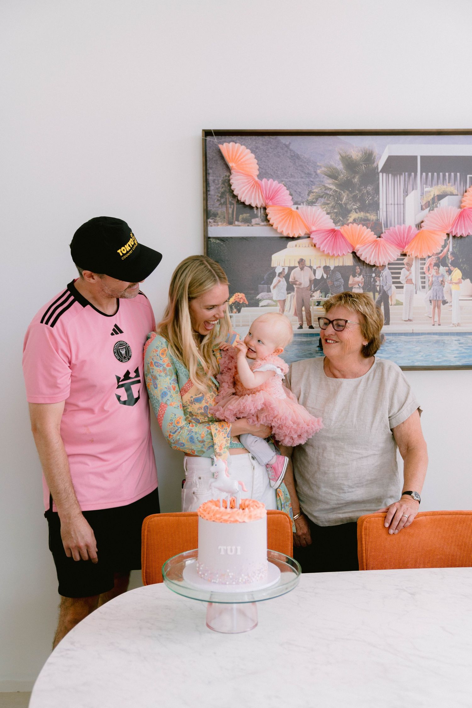 A pink-themed birthday celebration with a cake, decorative paper fans, and family gathering around a white table.
