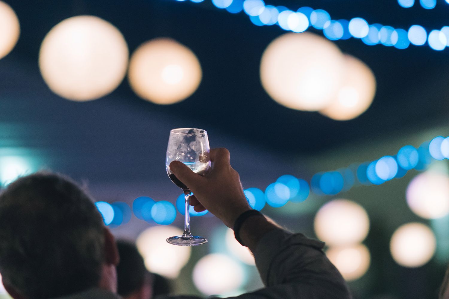 Hand raises a wine glass against blurred bokeh lights in evening atmosphere.