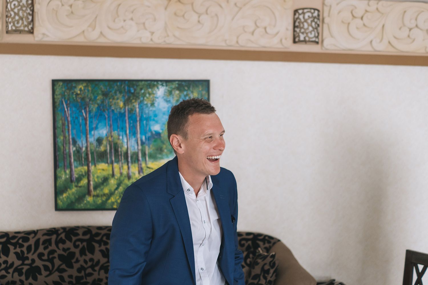 Person in navy blazer and white shirt stands near a landscape painting smiling.