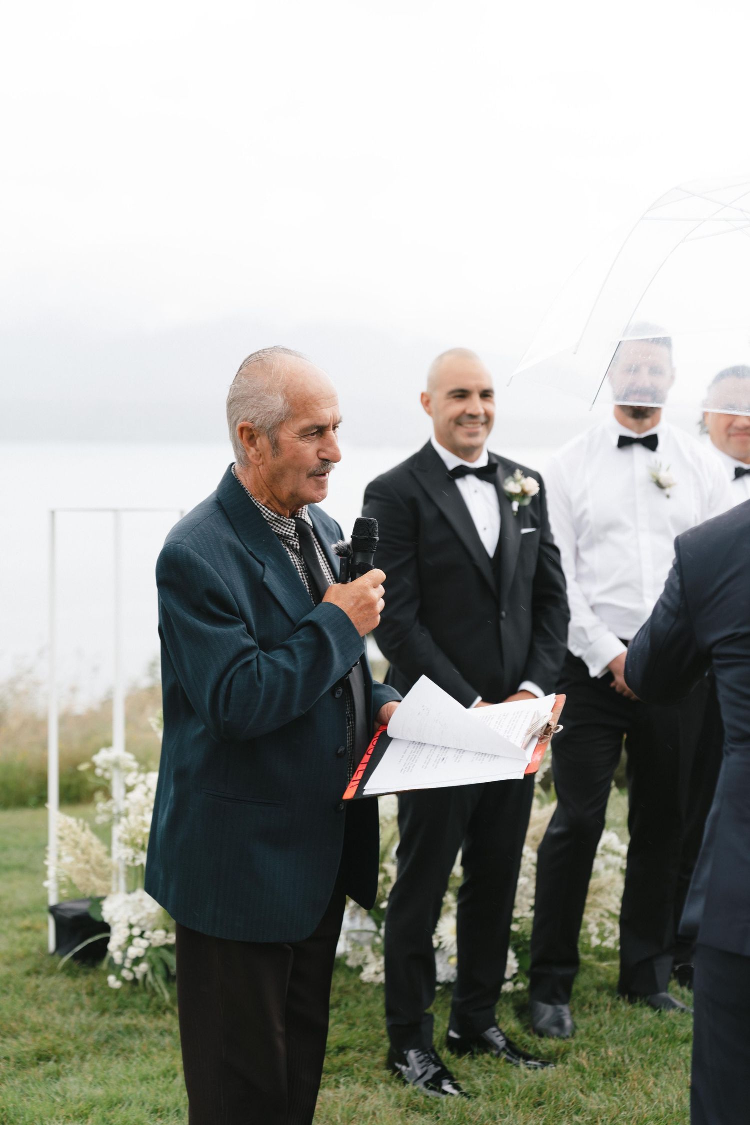 An outdoor wedding ceremony with an officiant speaking into a microphone as attendees gather on grassy terrain.