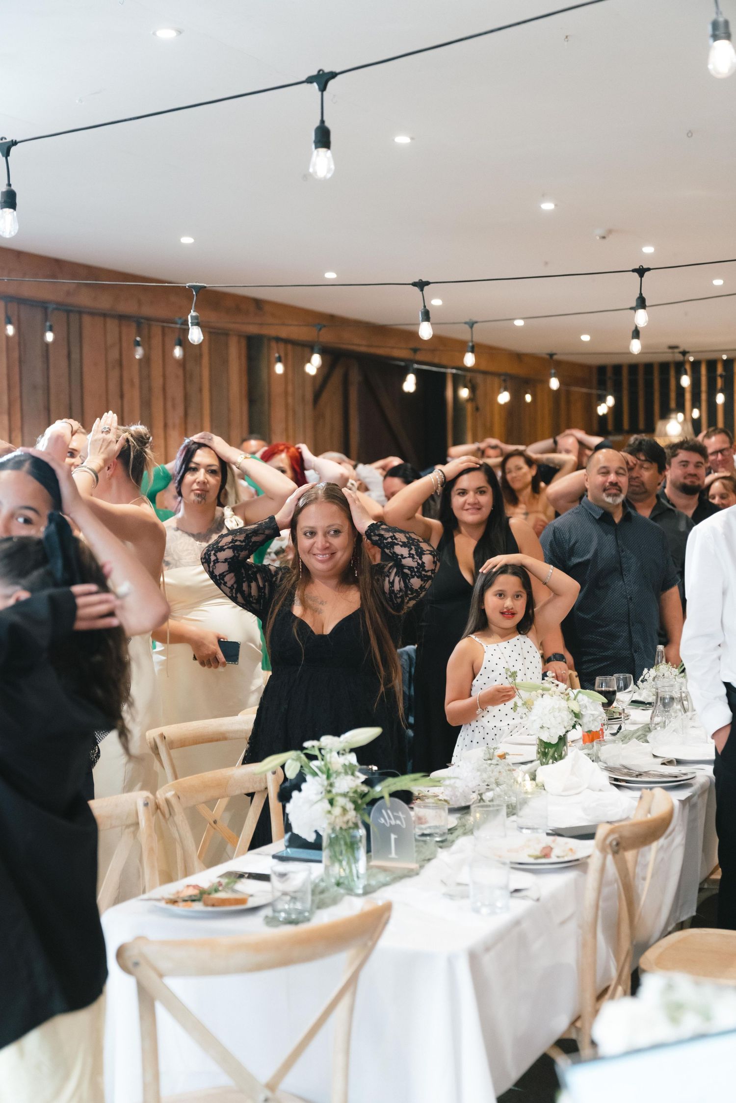 Wedding reception guests perform synchronized dance movements at elegant tables decorated with white linens and floral centerpieces.