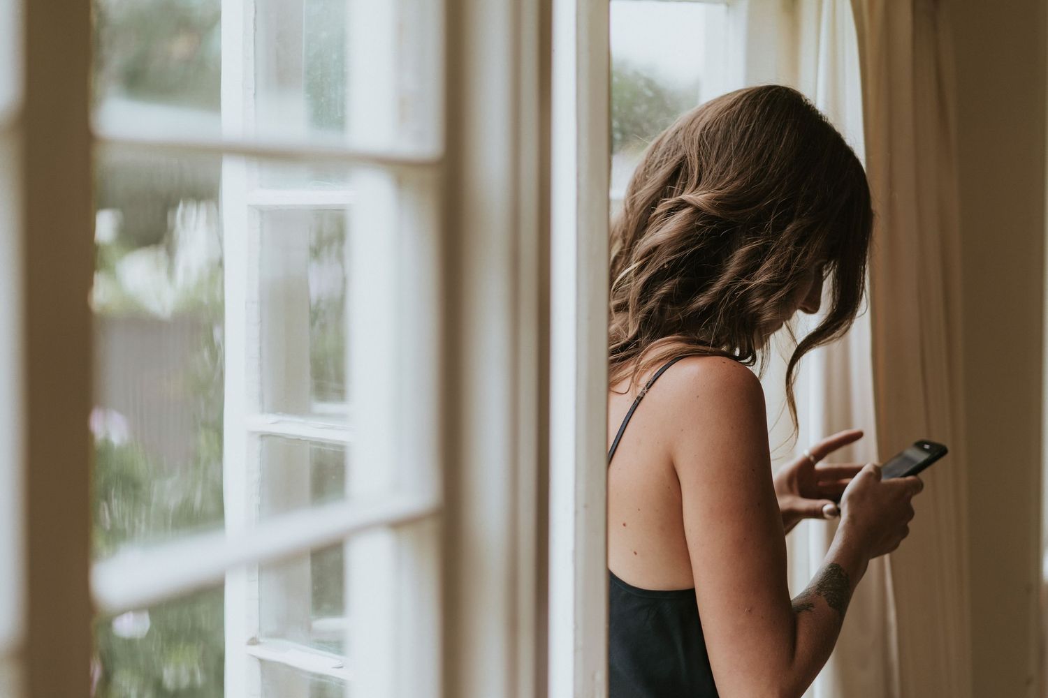 A person in a black tank top stands by a window looking at their mobile phone in natural light.