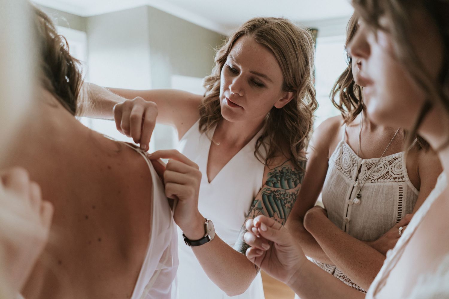Wedding preparations underway as people help adjust a white dress in a soft, natural light filled setting.