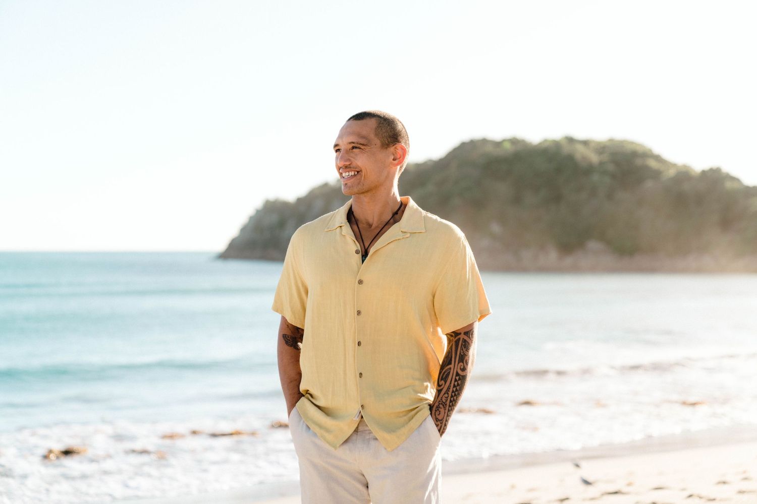 Person in yellow shirt with hands in pockets standing on serene beach with coastal cliffs in background.