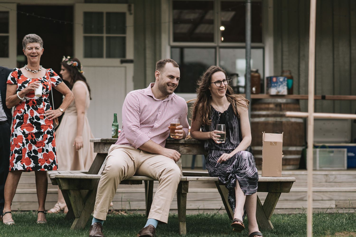 People enjoying drinks and conversation while sitting on wooden benches at an outdoor gathering.
