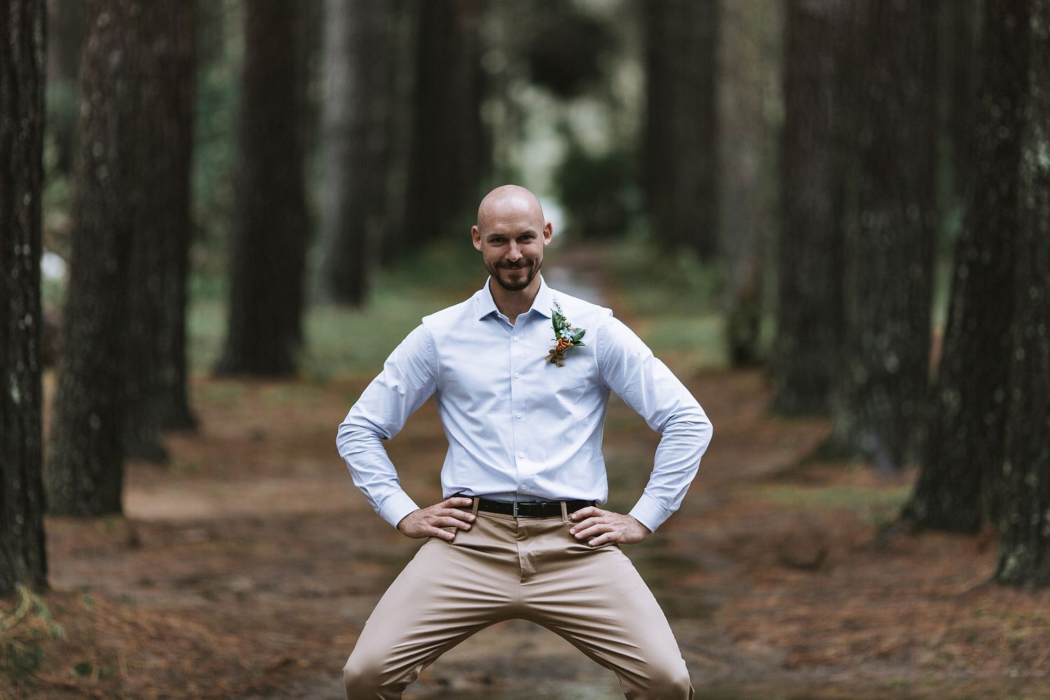 A person in a white shirt and khaki pants strikes a playful pose on a forest trail surrounded by pine trees.