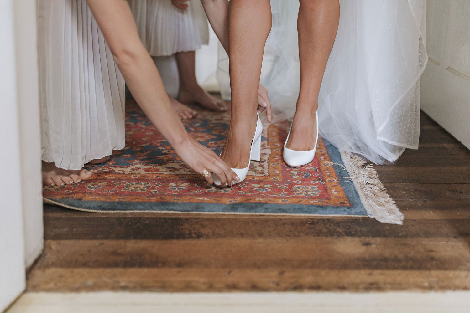 Two pairs of hands help put on white ballet flats and heels while standing on a vintage rug at a doorway.