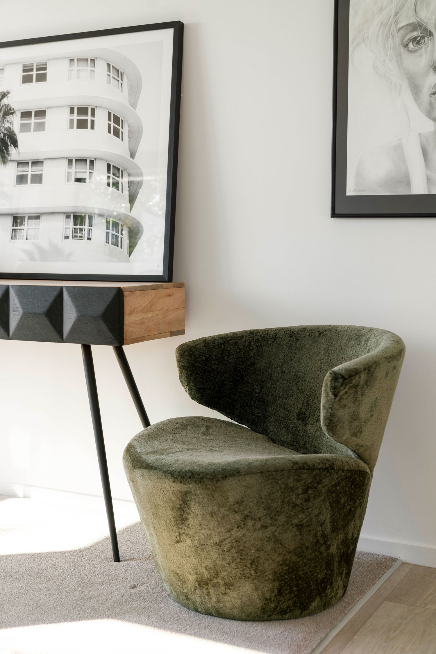 A curved velvet accent chair in olive green is displayed against a white wall with framed artwork.