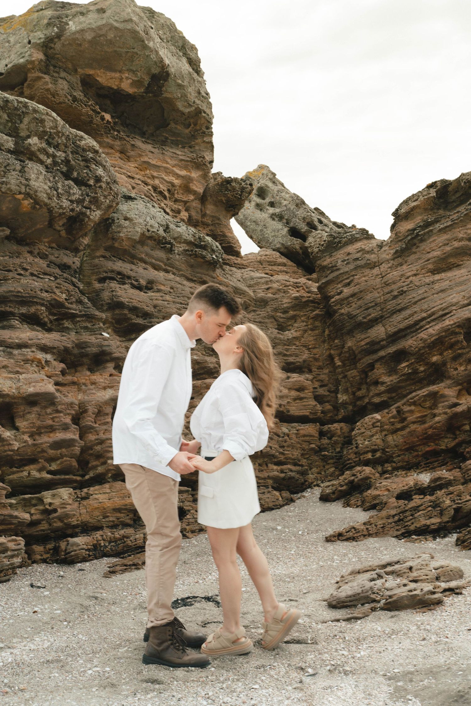 A couple in matching white outfits share an intimate moment among dramatic coastal rock formations.