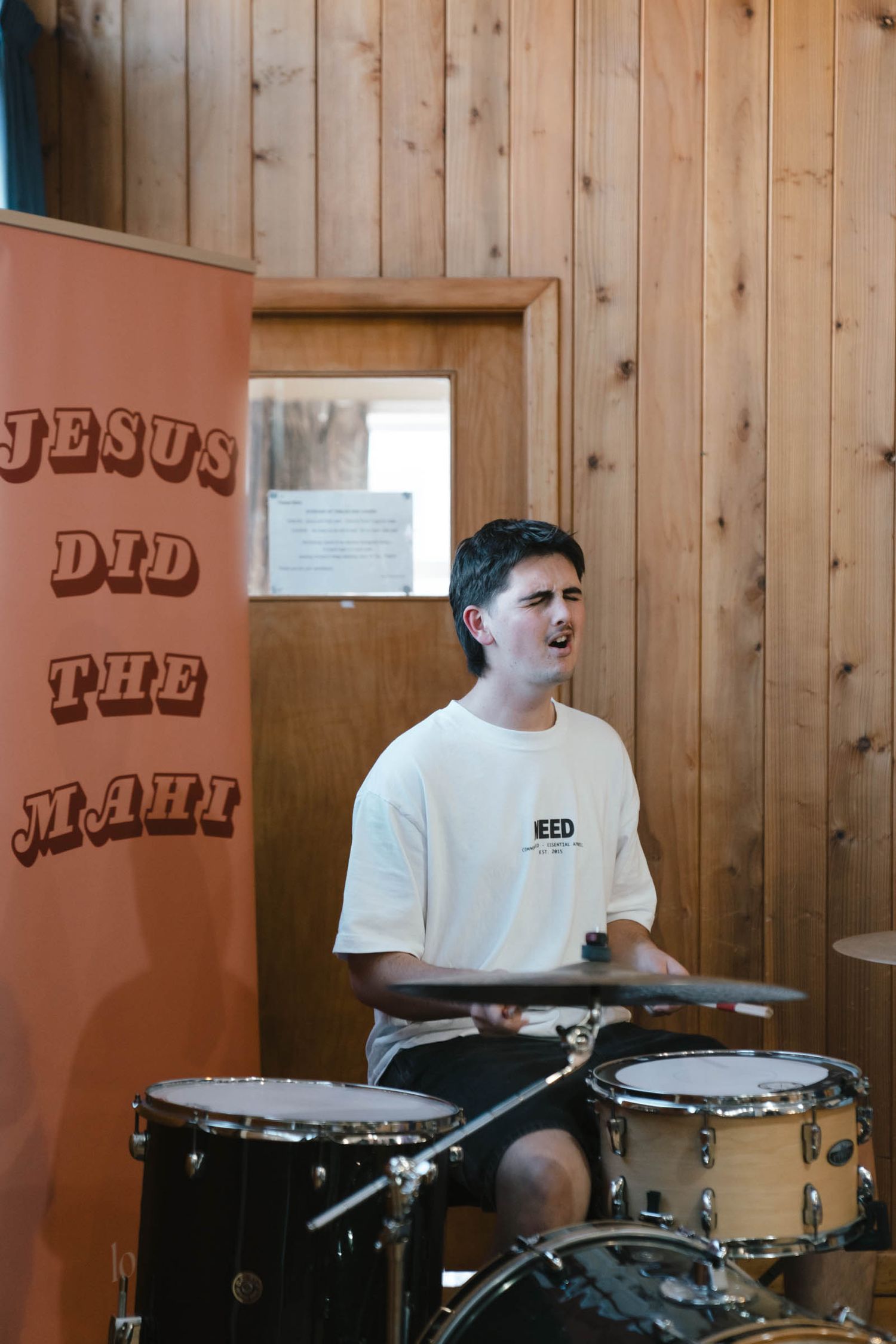 A drummer performing on stage with 'Jesus did the math' text visible on the wall.