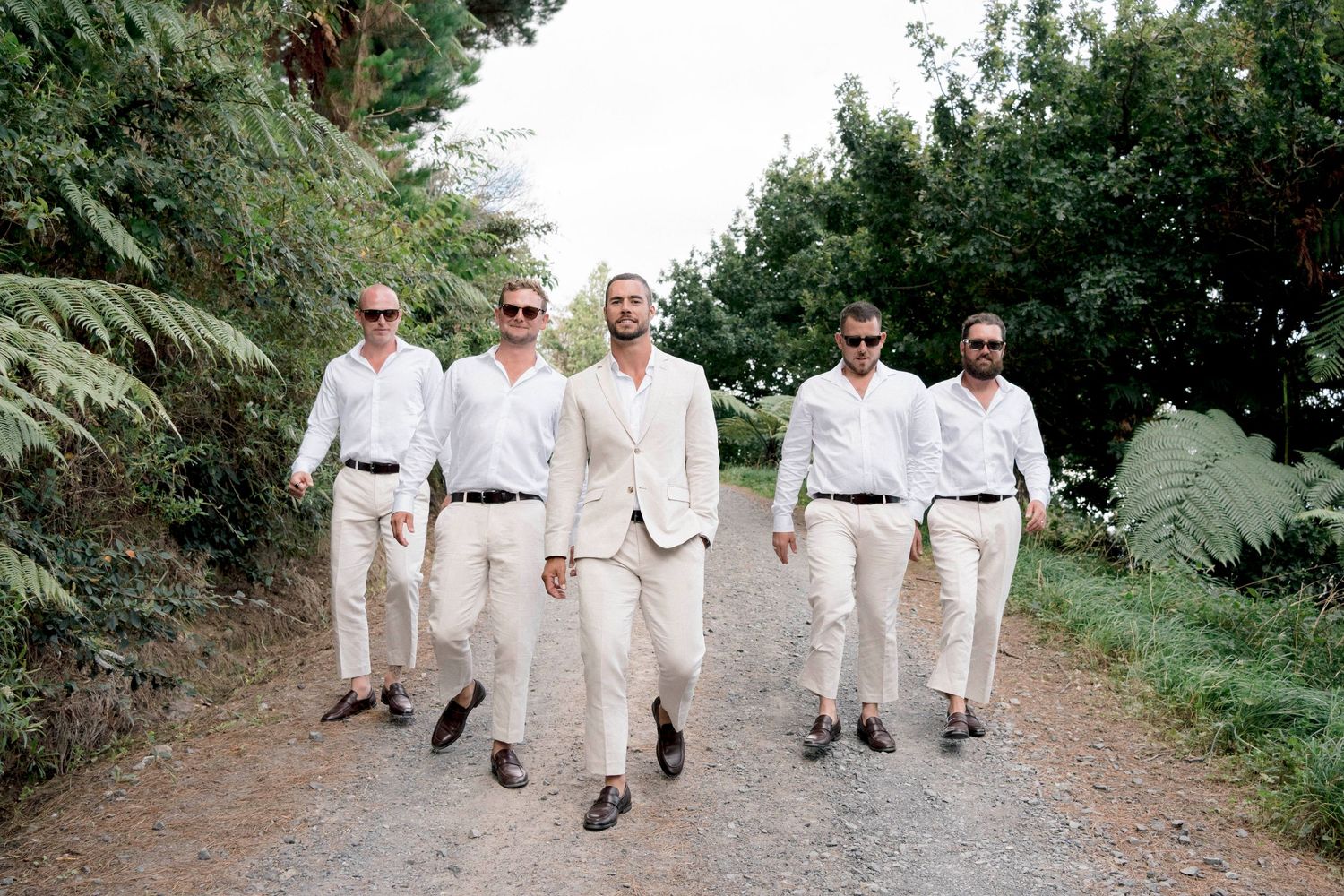 Groomsmen in coordinated cream and white outfits walking down a garden path.