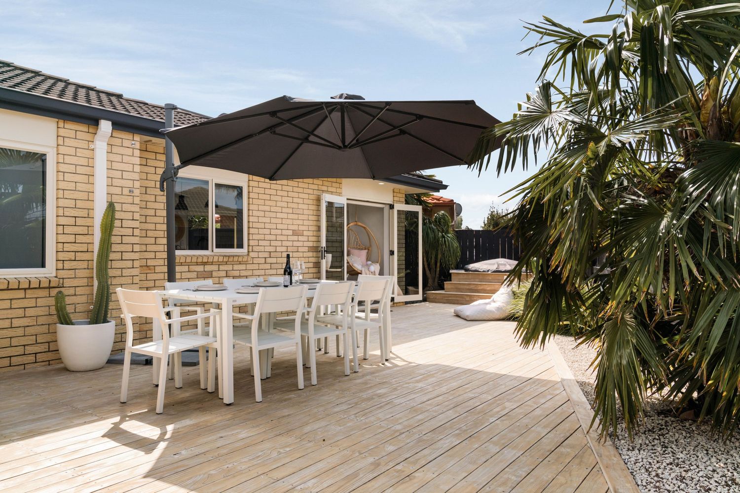 Beige brick house exterior with white outdoor dining furniture and umbrella on wooden deck near palm tree.