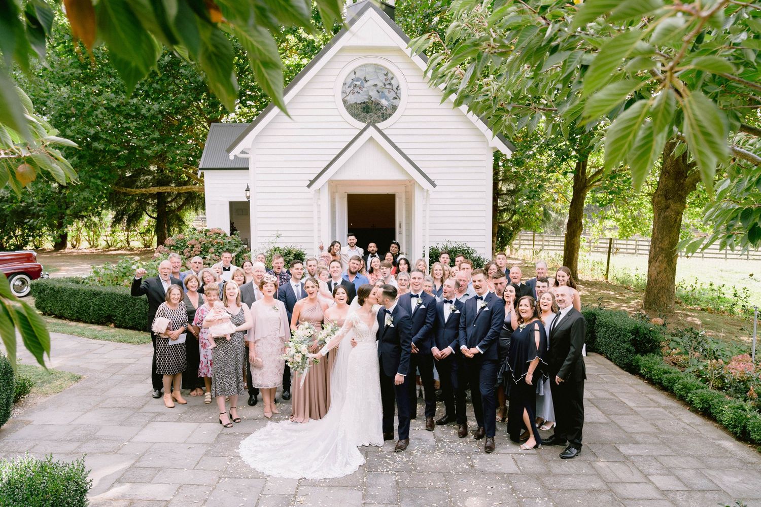 Wedding party gathered outside white chapel with tree-lined walkway on sunny summer day.