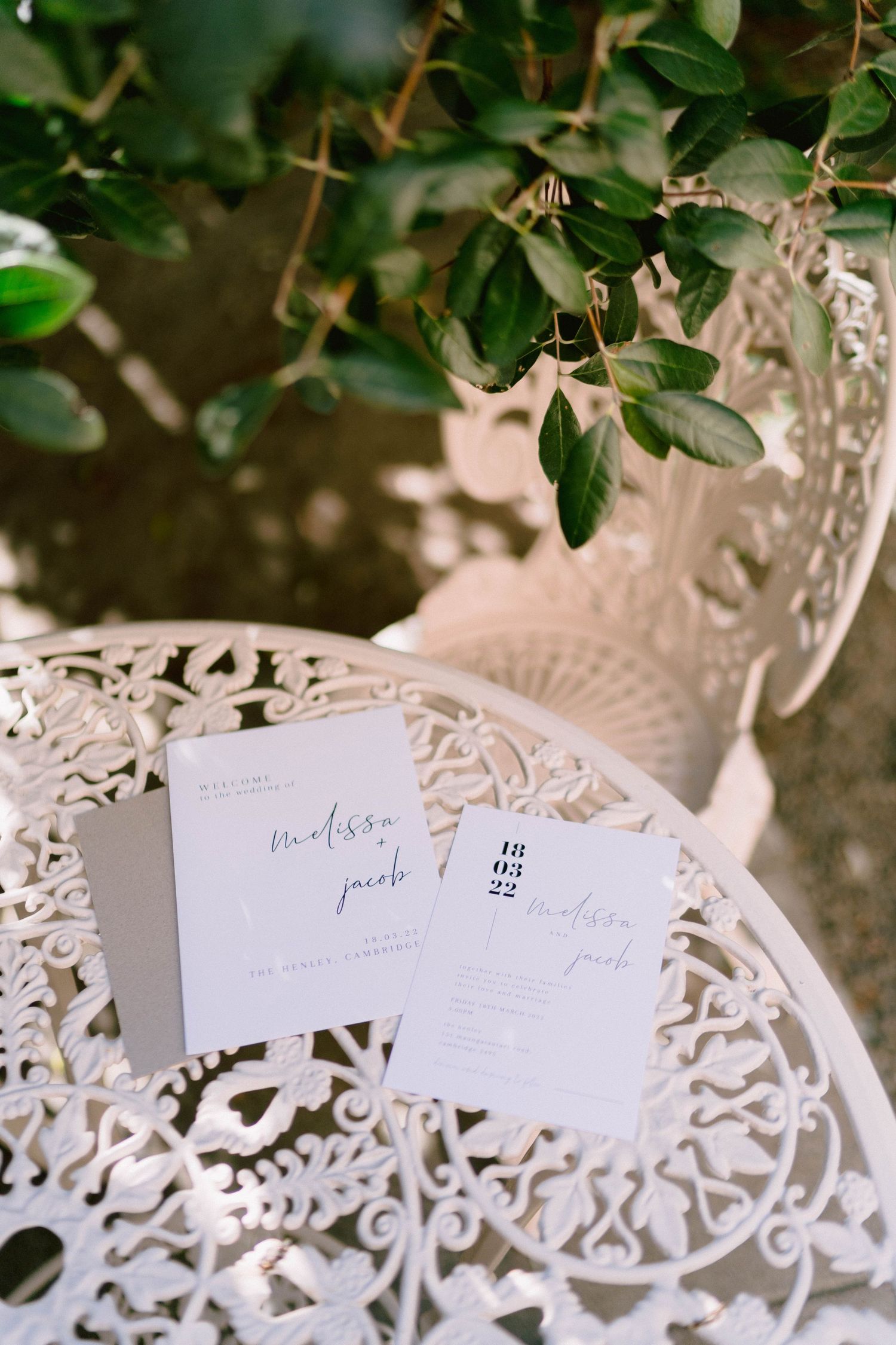 Wedding invitation cards displayed on an ornate white metal garden chair with green foliage in the background.