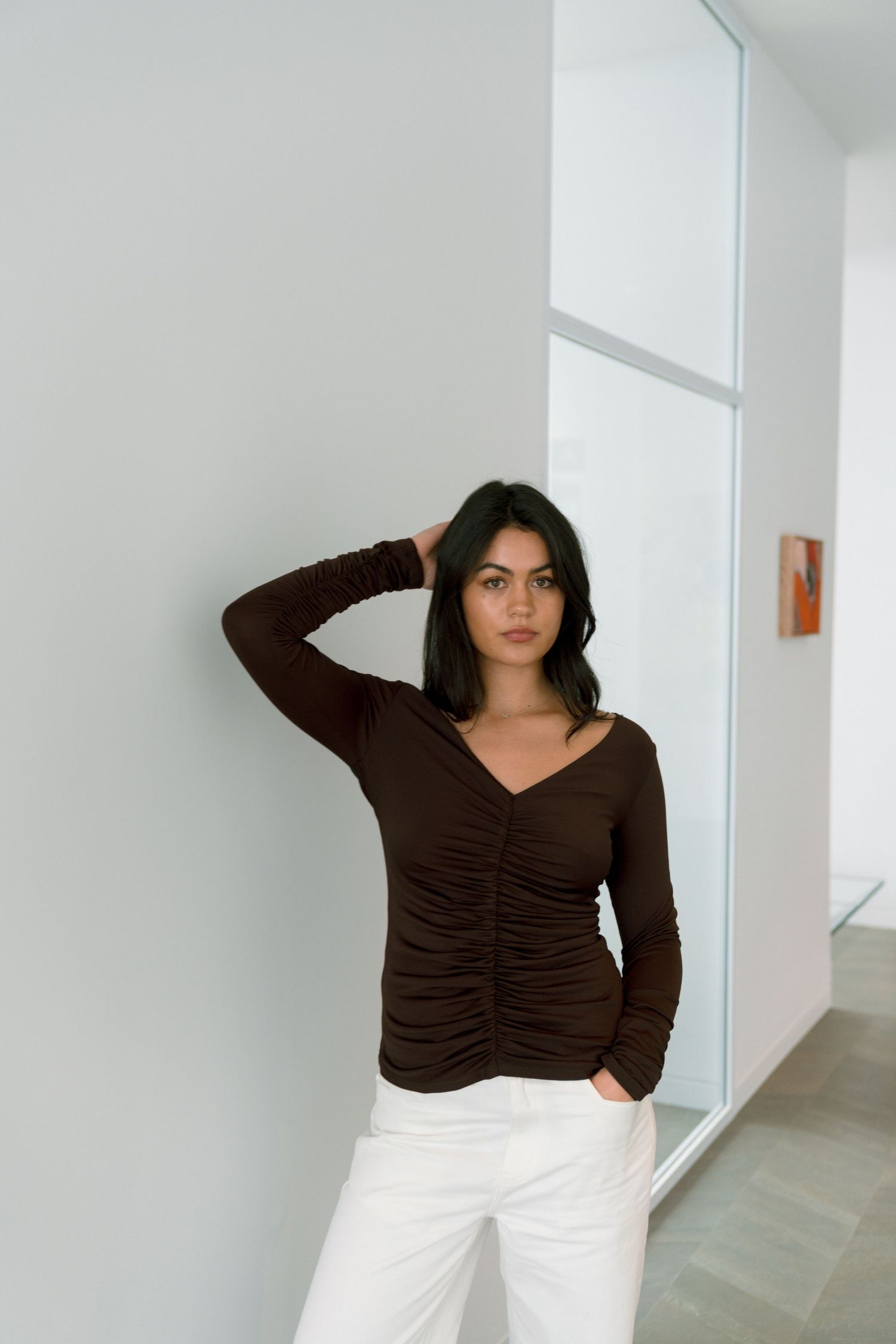 Woman in brown ruched long-sleeve top and white pants leaning against a white wall.