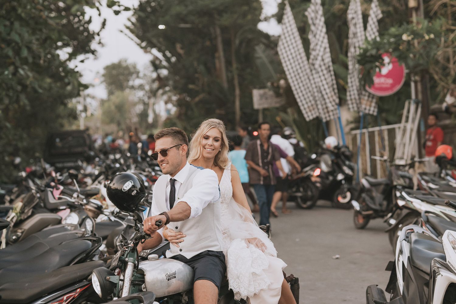 Bride and groom posing with motorcycles parked along a tree-lined street.
