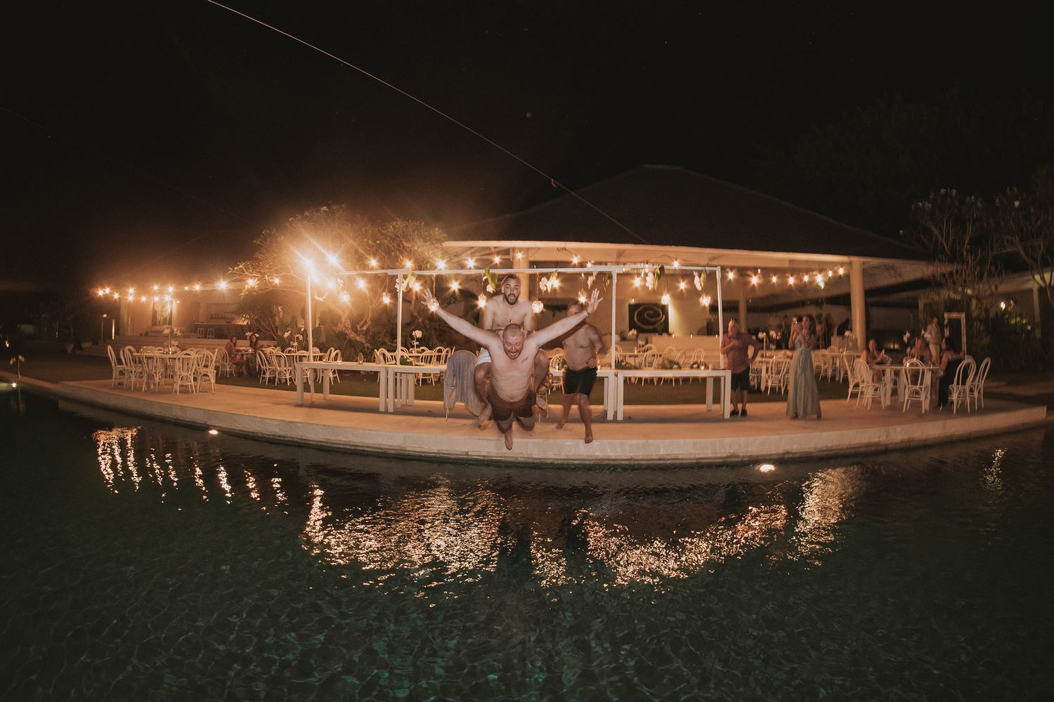 Panoramic view of a nighttime wedding reception with glowing lights and poolside setting.