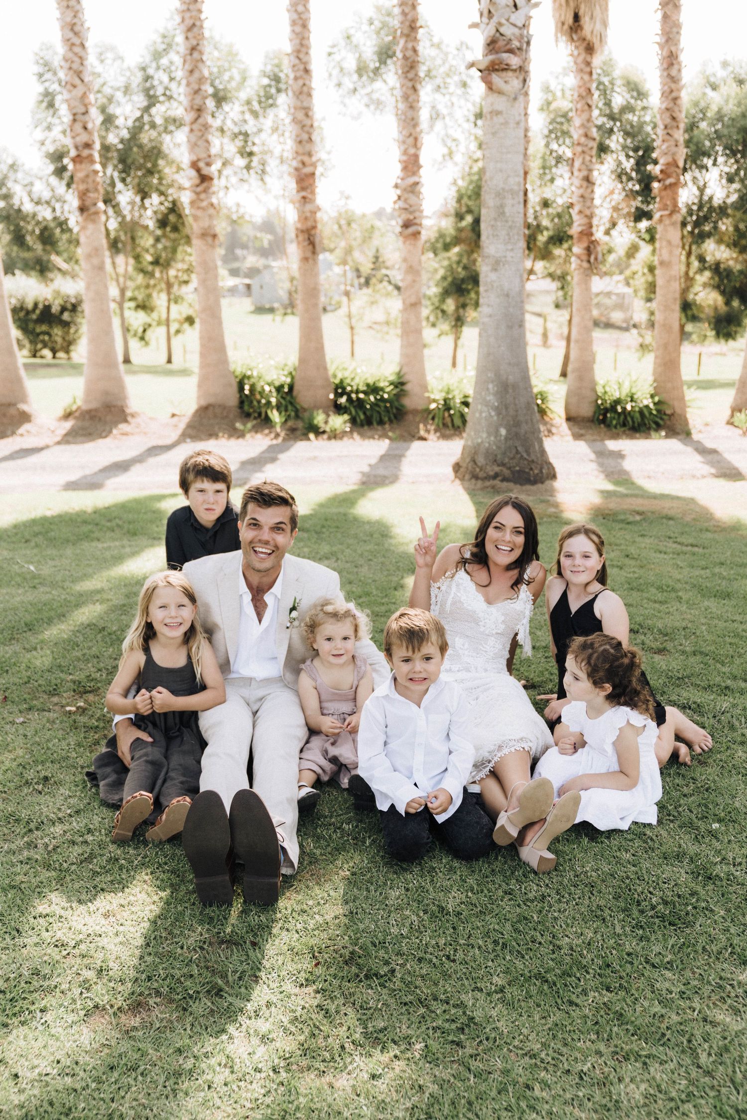 A group sits together on grass beneath tall palm trees in coordinated white outfits.