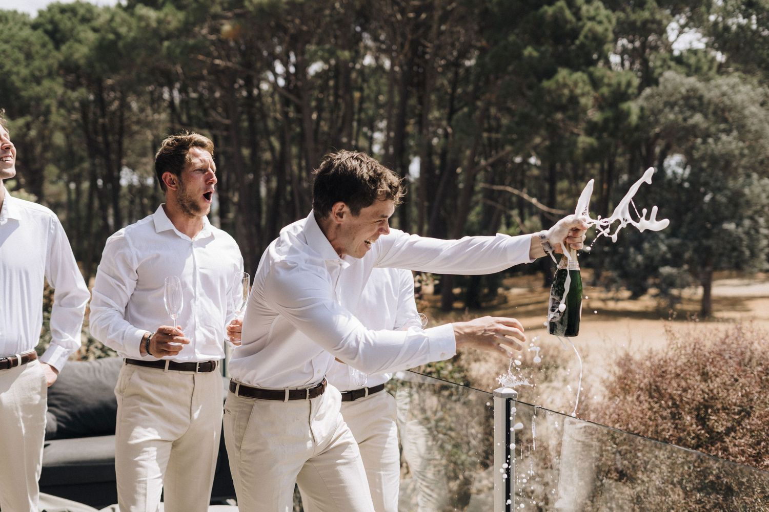 Group wearing white shirts gathered outdoors in a pine forest setting raising drinks in celebration.
