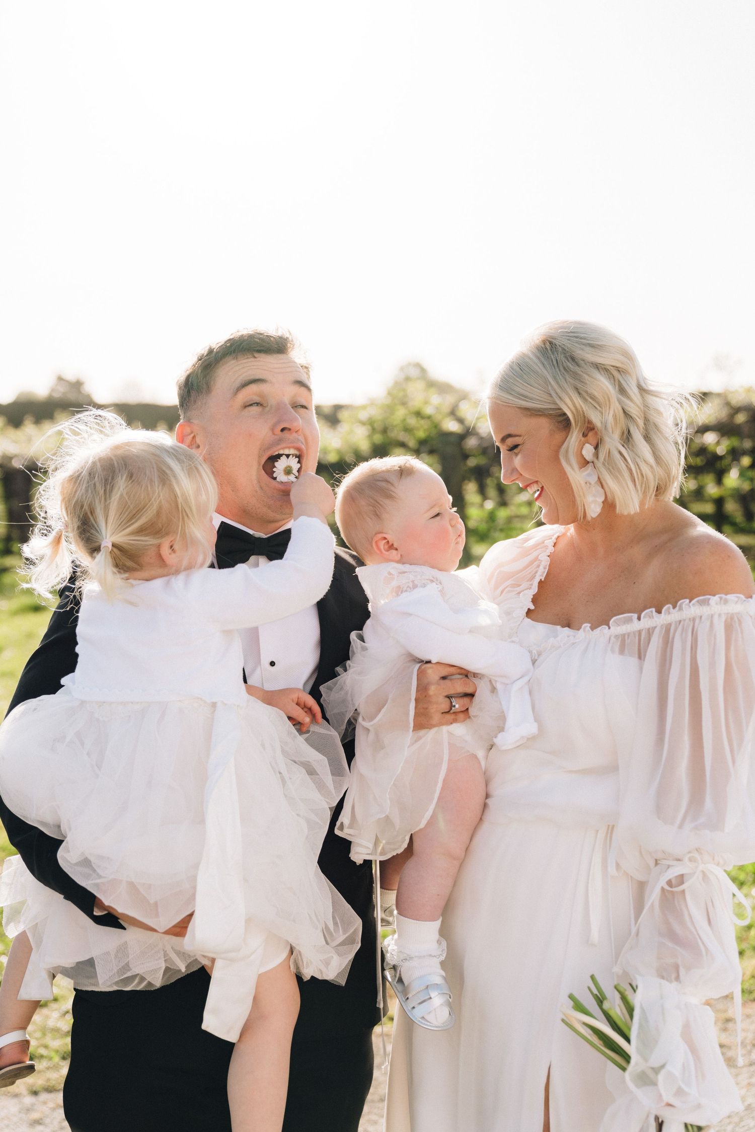 Joyful wedding portrait showing couple in formal attire with small children in white dresses enjoying outdoor celebration.