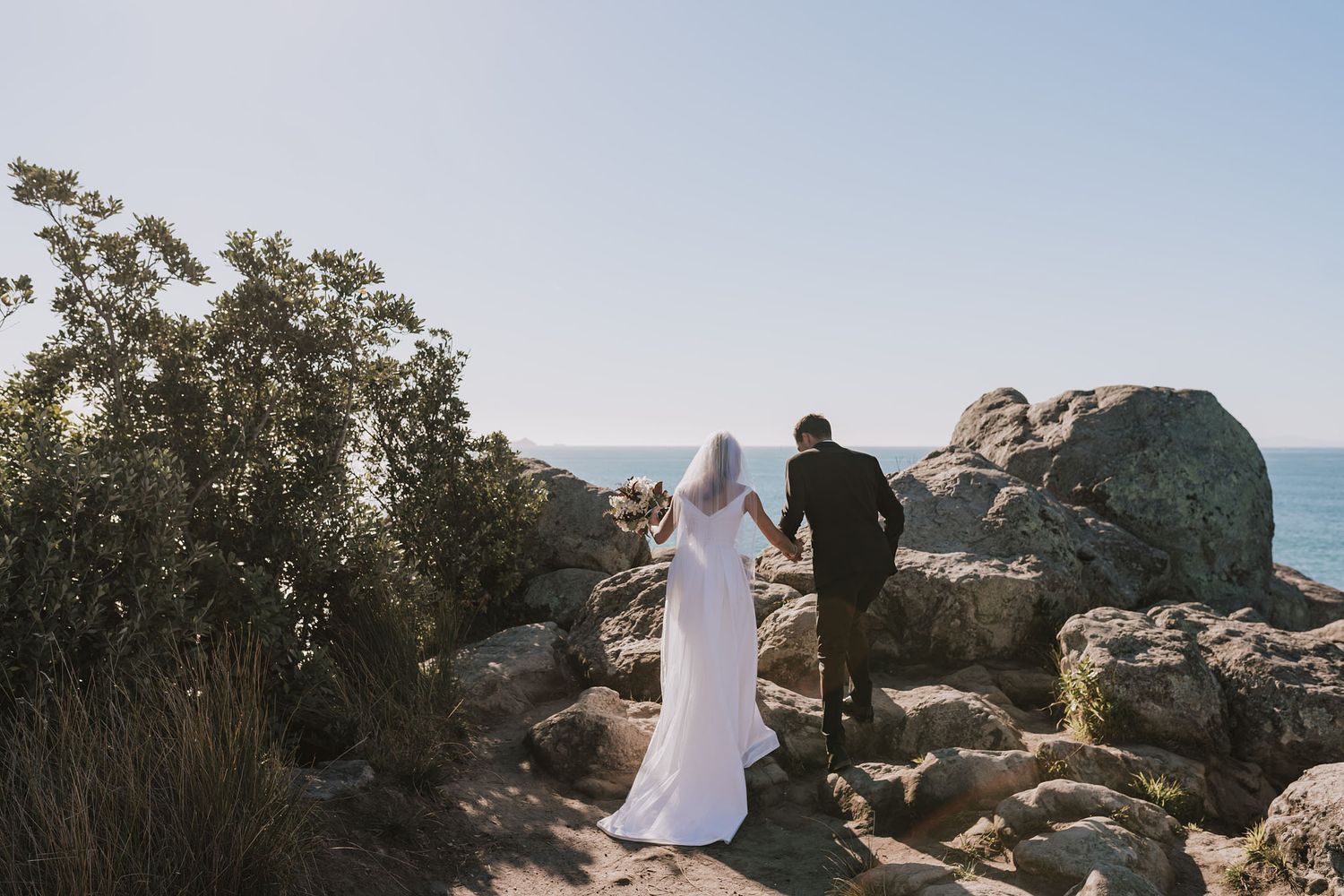 A couple stands together on rocky coastal cliffs overlooking the ocean against a bright blue sky.