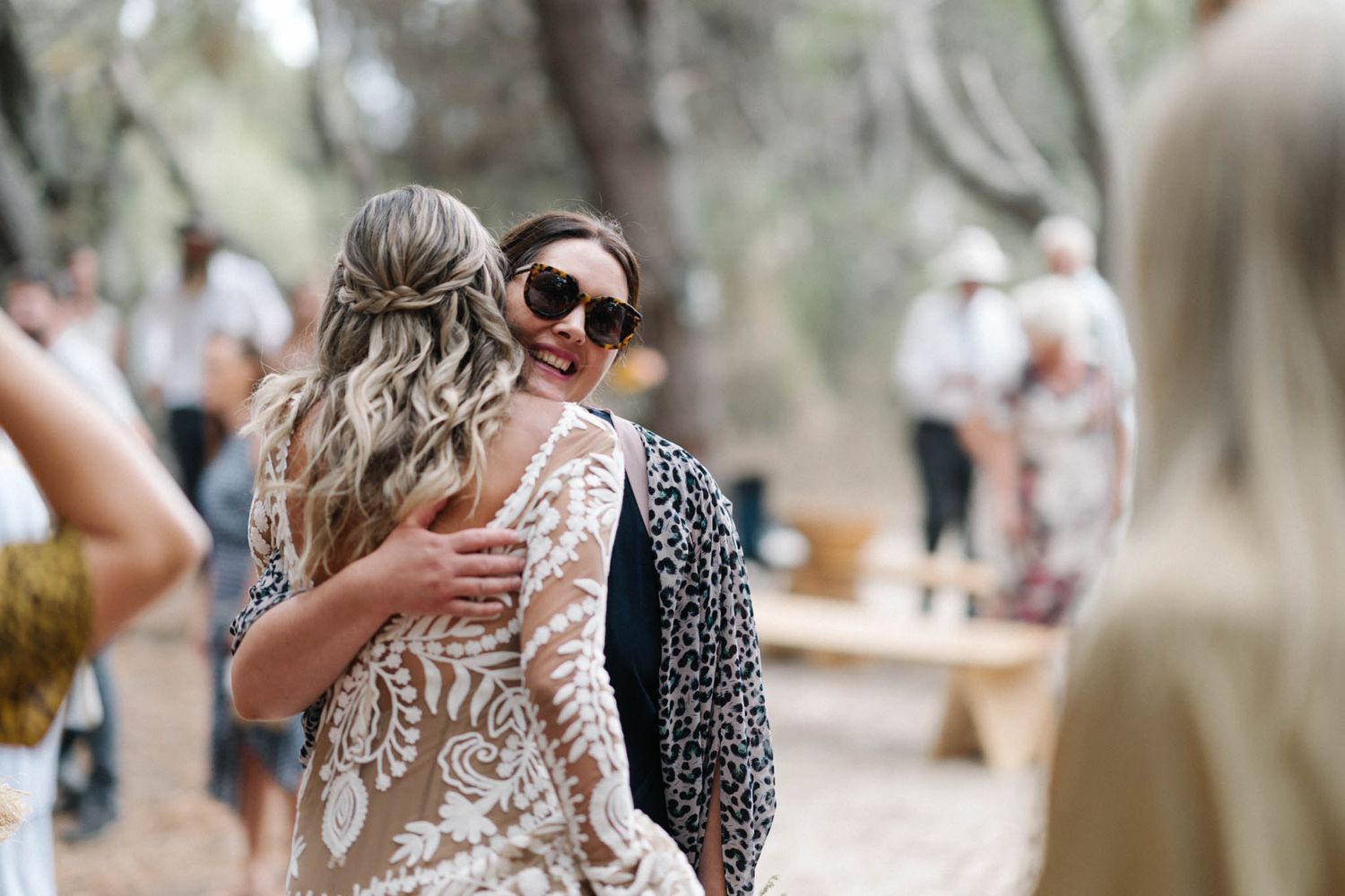 Wedding guests enjoy outdoor celebration with drinks under rustic wooden string lights and natural scenery.
