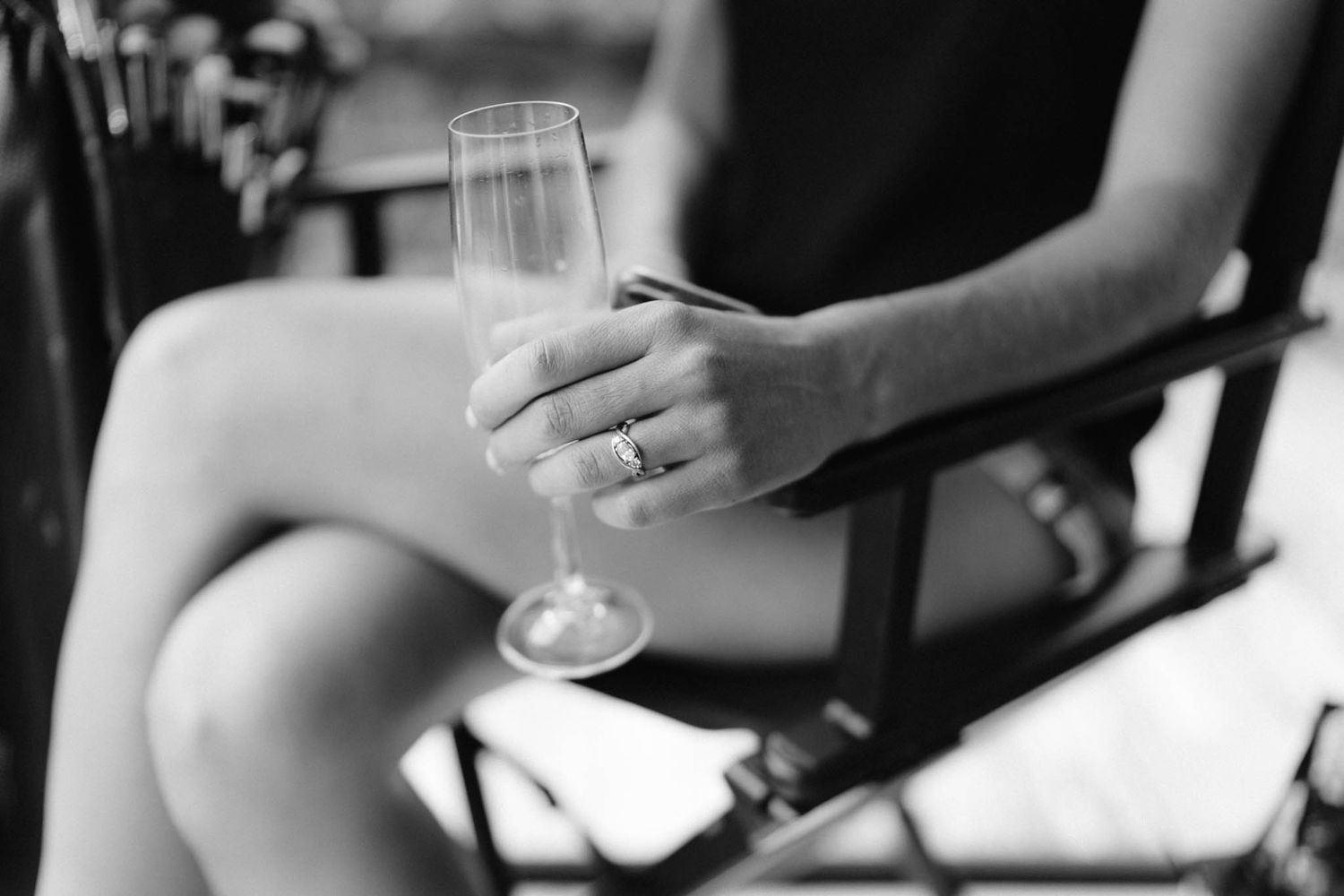 Black and white photo showing a hand holding a champagne flute while seated, with rings visible on fingers.