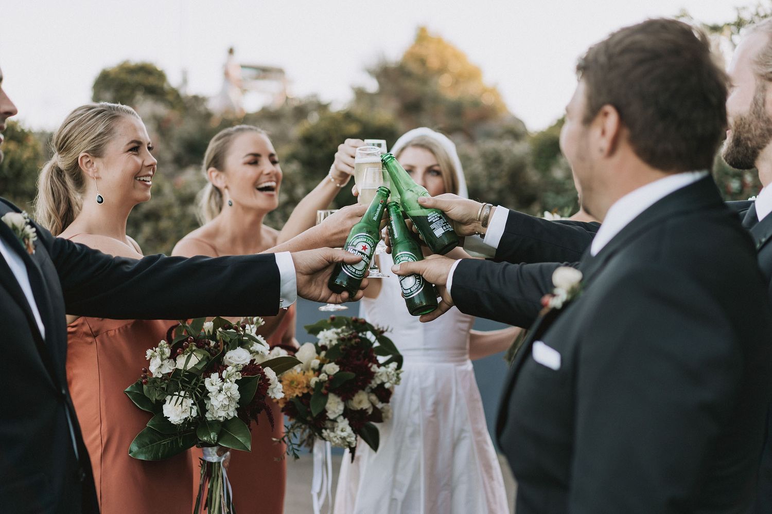 Wedding party toasting with glass bottles while celebrating at an outdoor venue.