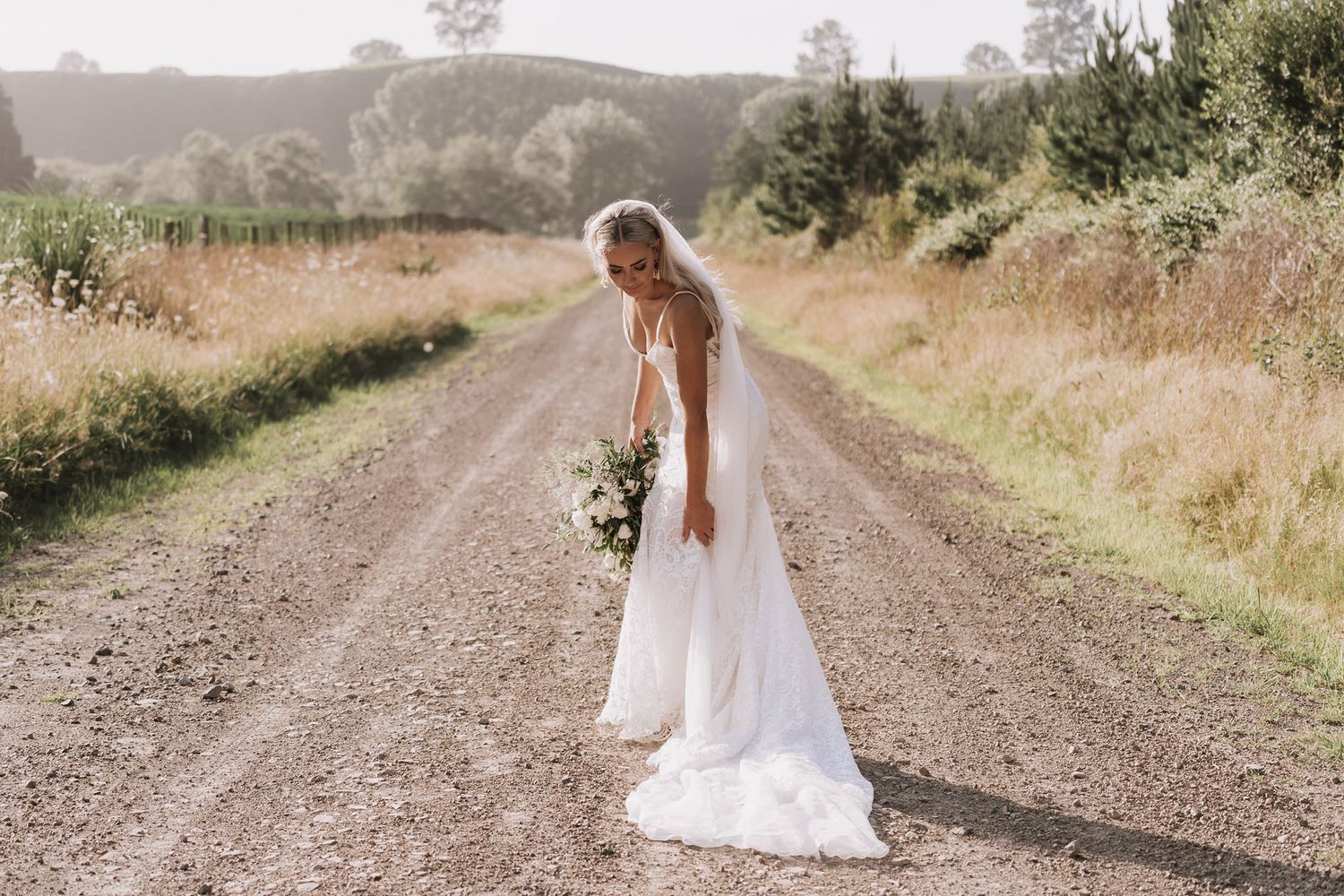 Person in flowing white dress walks down a serene dirt country road surrounded by golden grass and hills in summer.