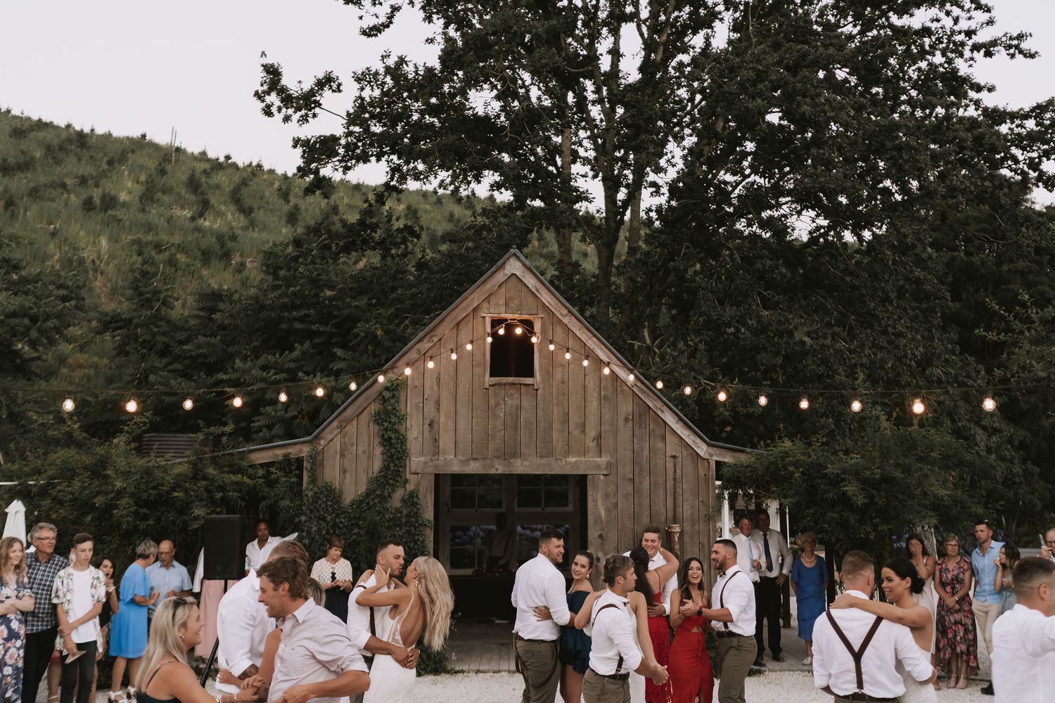 A rustic outdoor wedding ceremony with guests gathered under string lights near a wooden barn in the countryside.