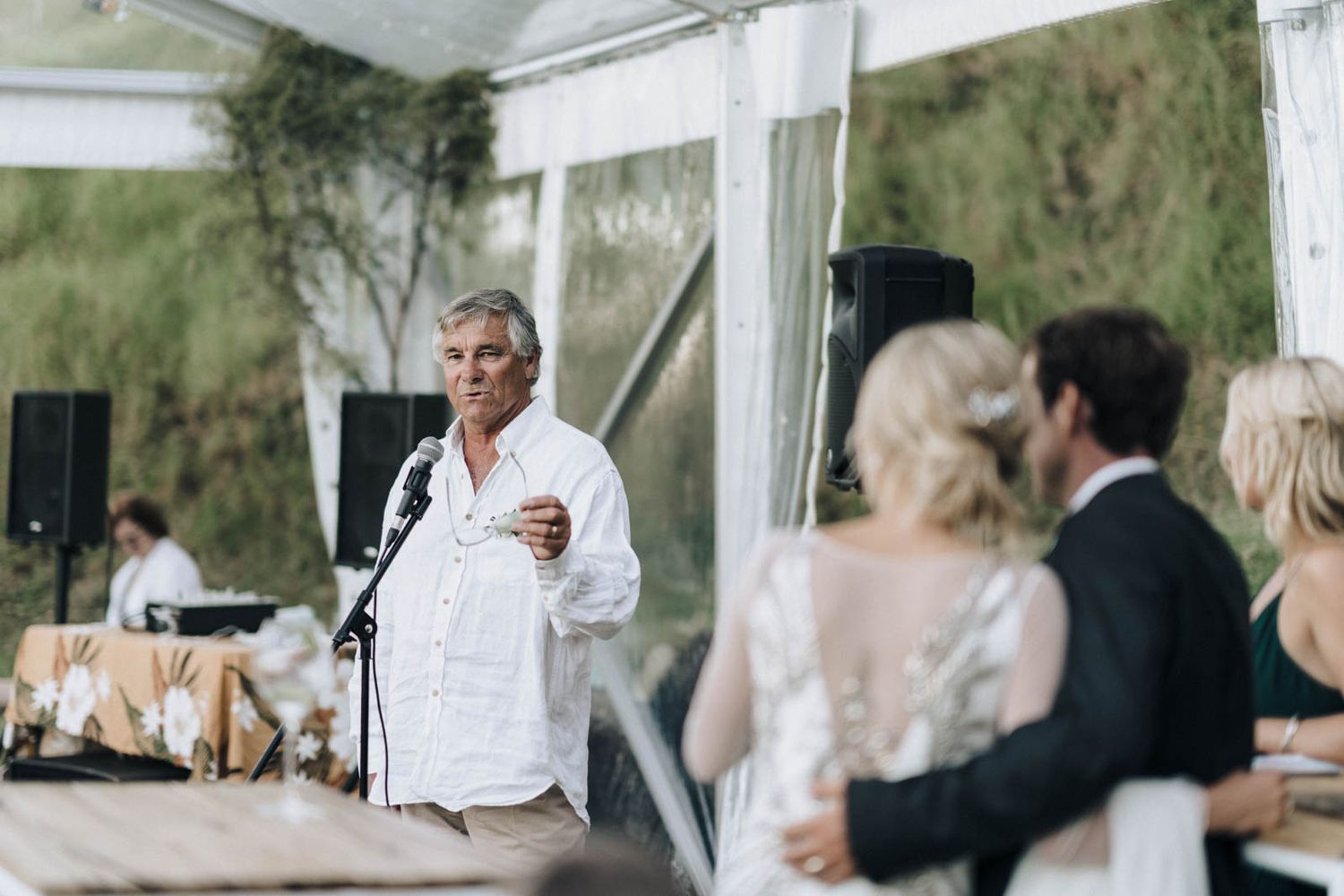 Wedding guests gather outdoors on a sunny day, with guests mingling and embracing while dressed in formal attire.