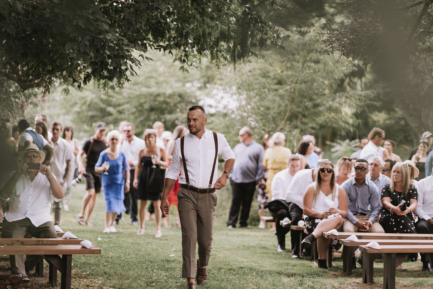 A rustic outdoor wedding ceremony in a garden setting with wooden benches and guests gathered under trees.