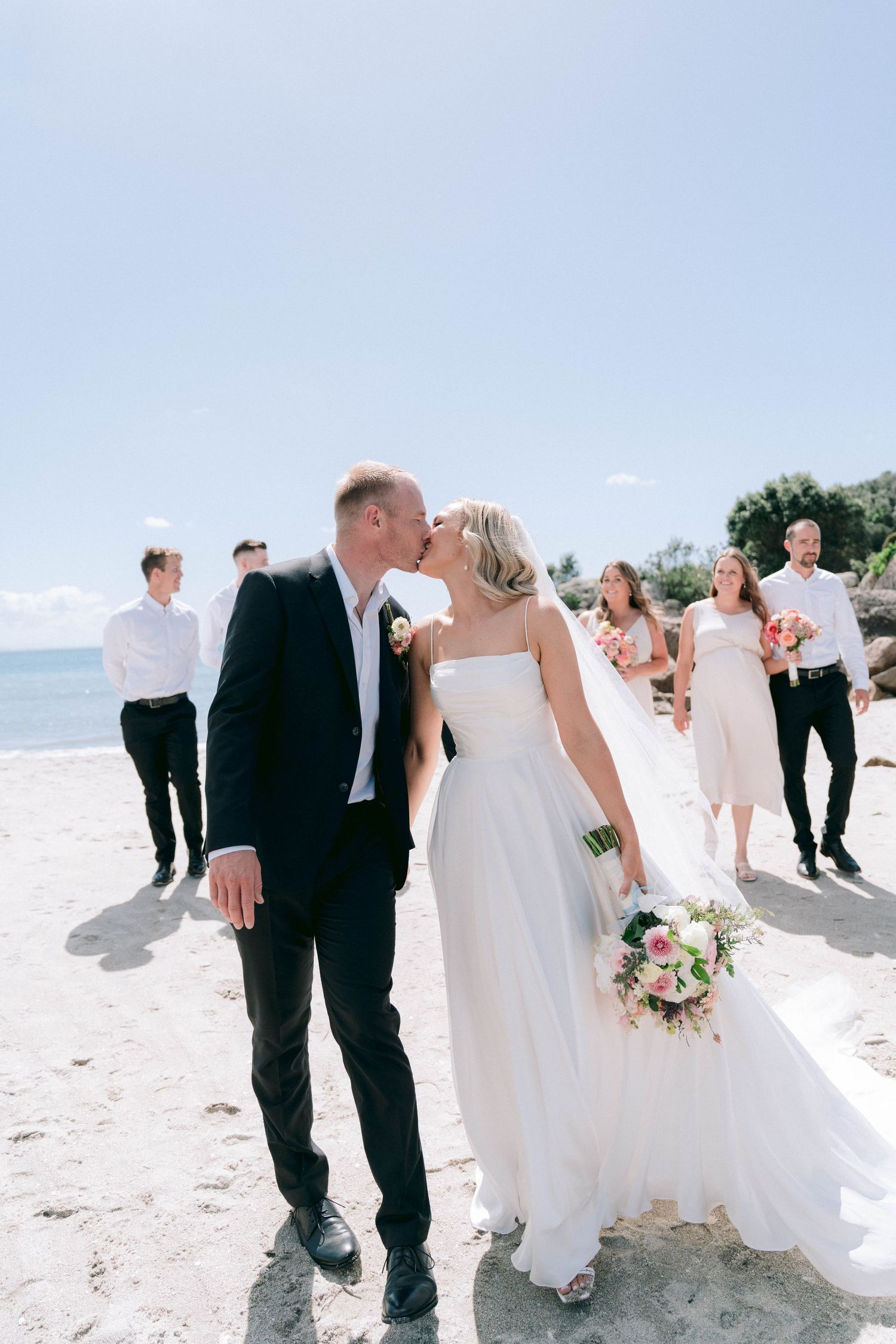 A romantic beach wedding ceremony with wedding party members in black and blush pink attire on a sunny day.