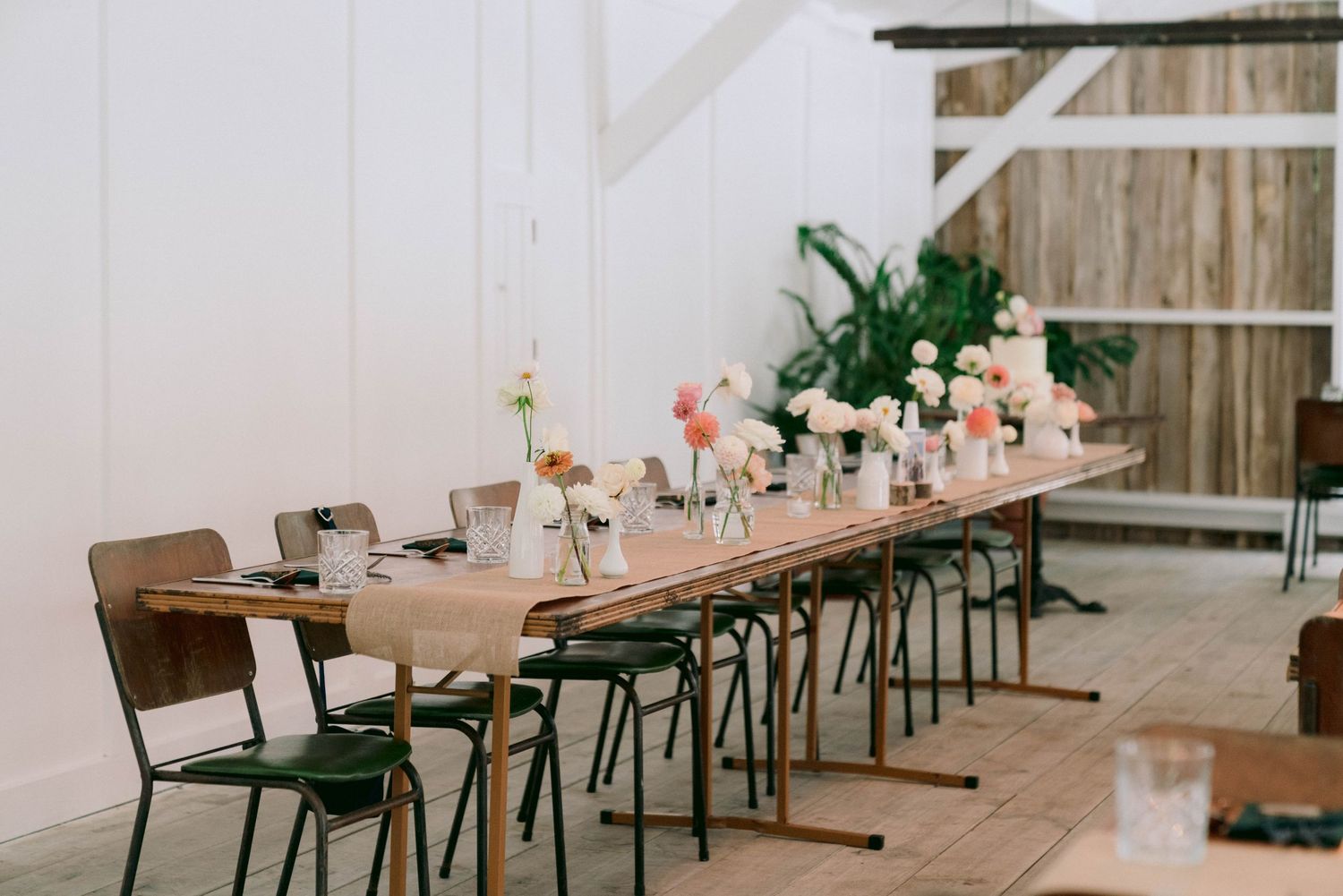 Long rustic dining tables with light wooden chairs and soft pink floral centerpieces create a romantic wedding reception setup.