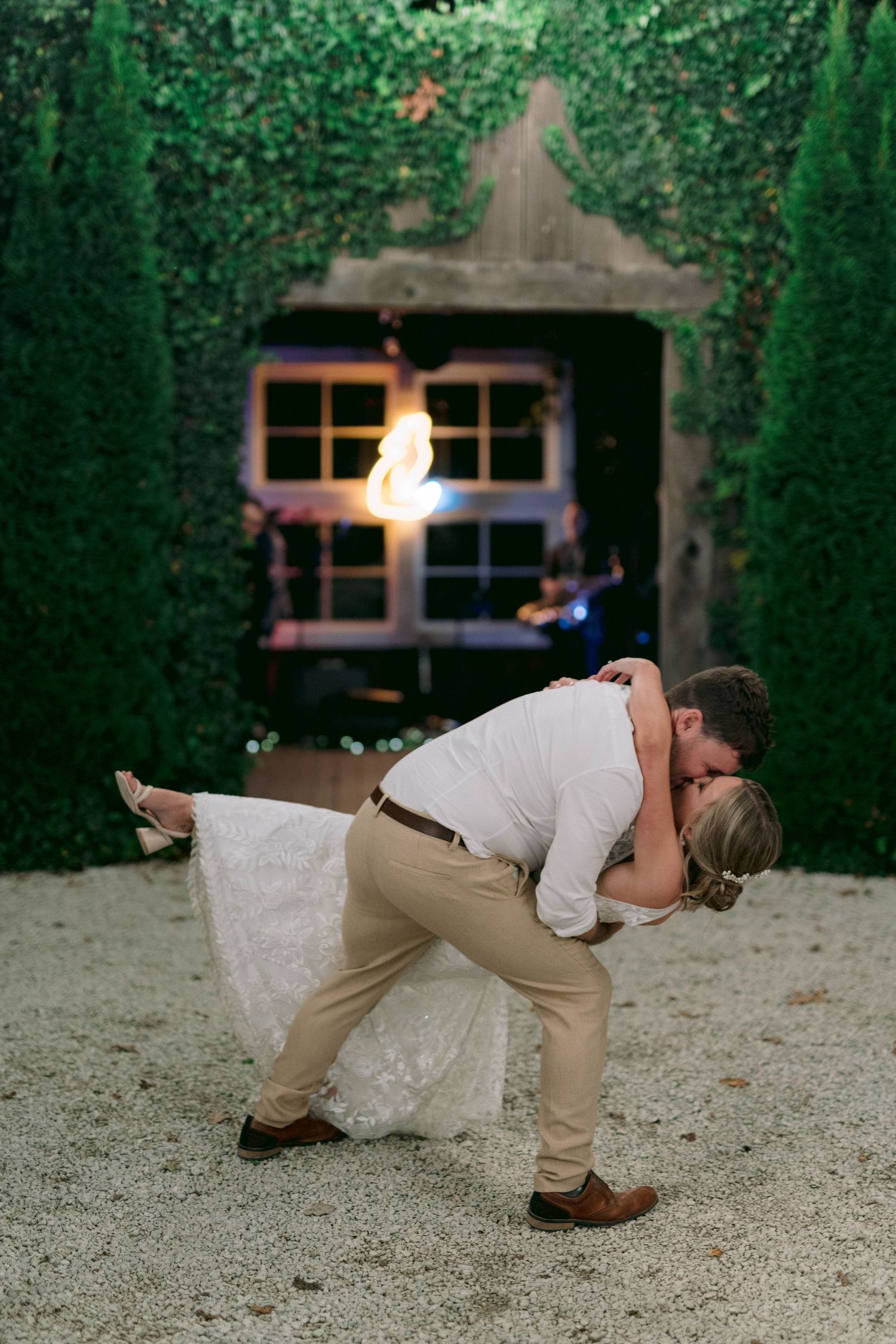 A romantic dip kiss between newlyweds against a green hedge wall with illuminated window at dusk.