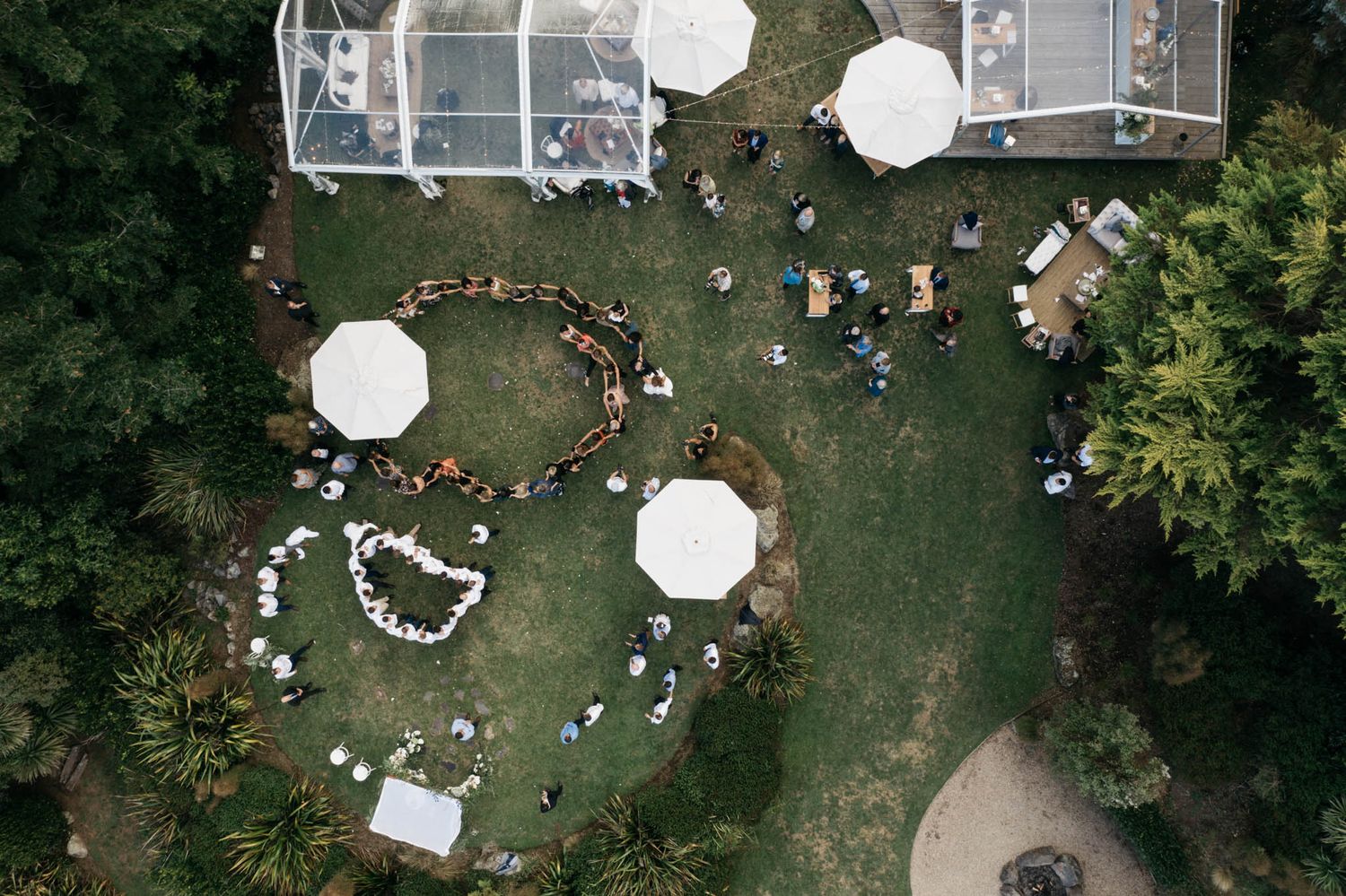 Aerial view of an outdoor wedding reception with white tents and guests arranged in heart-shaped seating on green lawn.