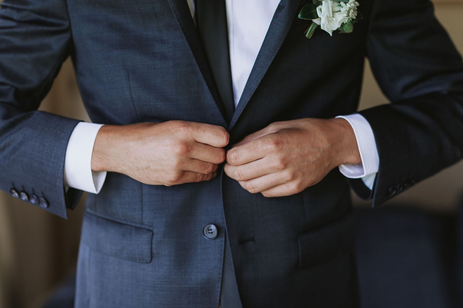 Close up detail of a groom adjusting his navy blue suit jacket with white boutonniere at formal wedding.