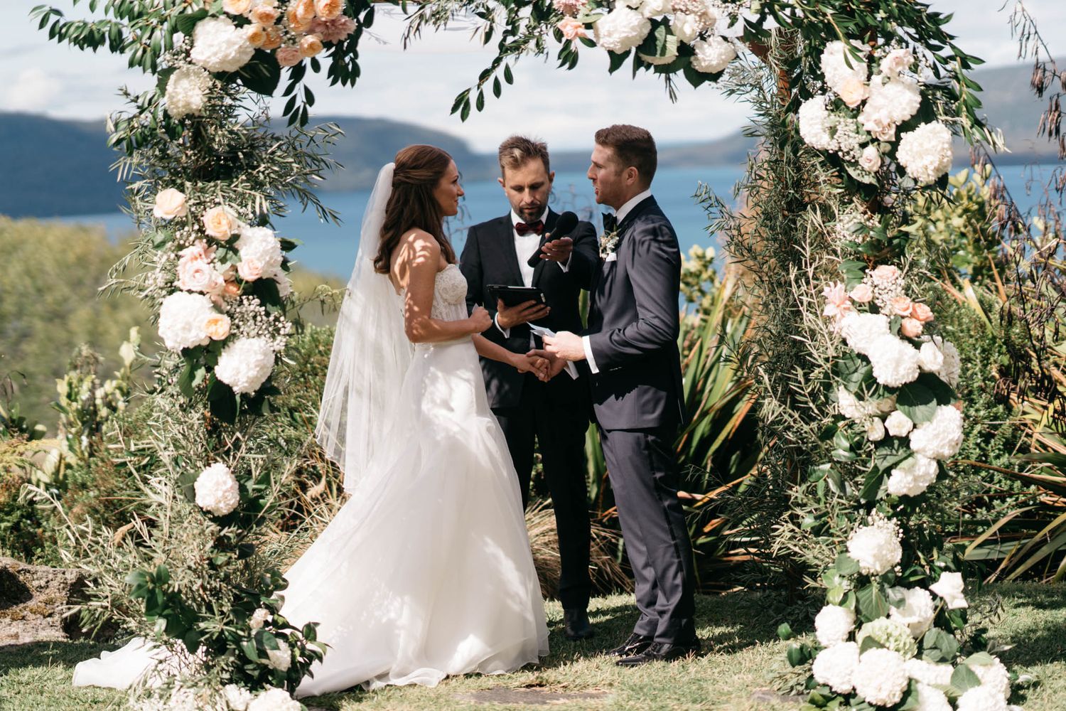 Outdoor wedding ceremony with white floral arch and mountain scenery in the background against a blue sky.