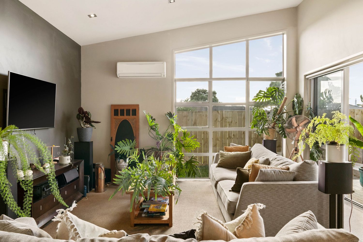 Modern living room with large windows, indoor plants, wall-mounted TV, and comfortable seating with neutral decor.