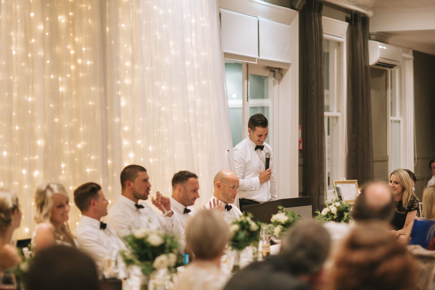 Best man delivers a wedding speech at an elegant reception with twinkling string lights and white floral centerpieces.