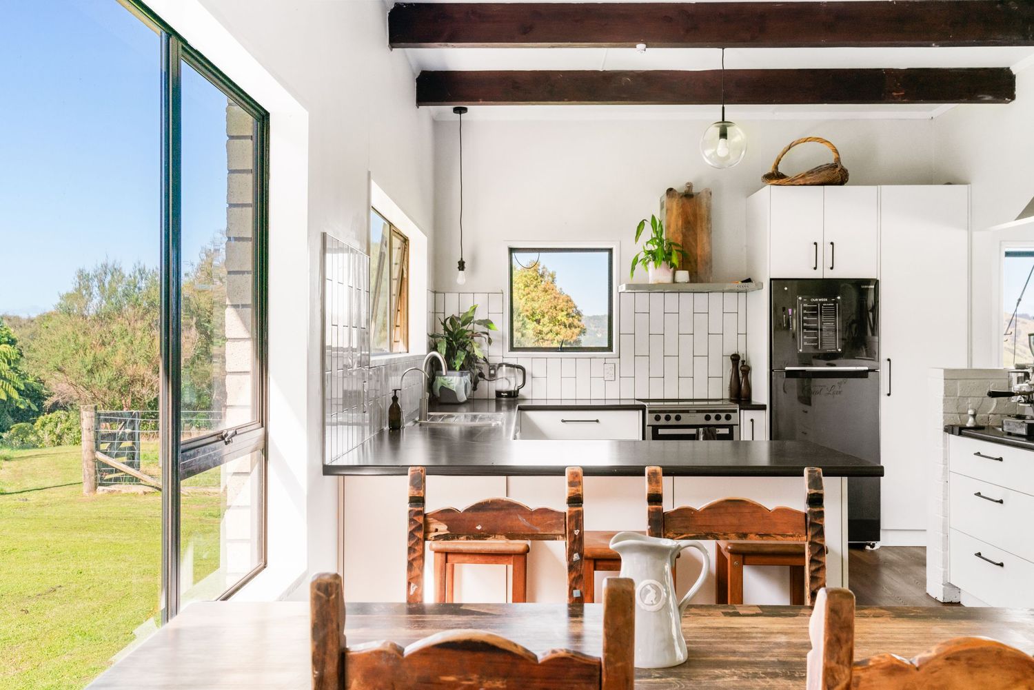 Modern white kitchen with exposed wooden beams, wooden dining chairs and large windows overlooking a sunny yard.