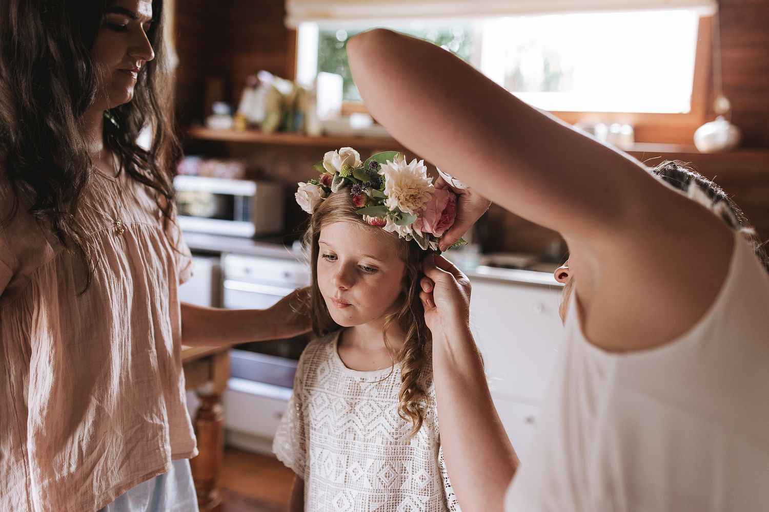 A person wearing a delicate floral crown and white lace dress is being helped with final touches in a rustic interior.