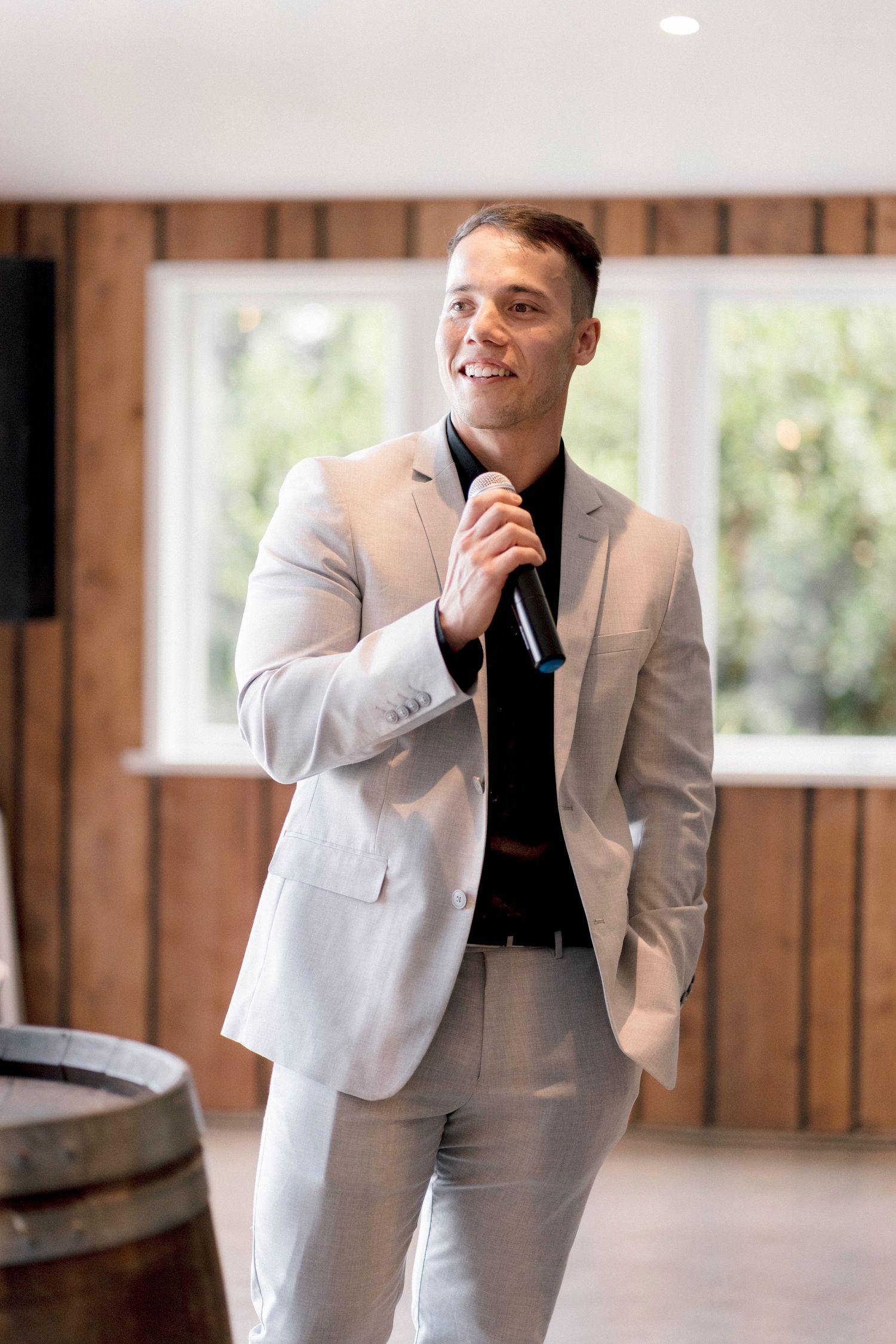 A person wearing a light gray suit and black tie speaks into a microphone in a rustic indoor venue.