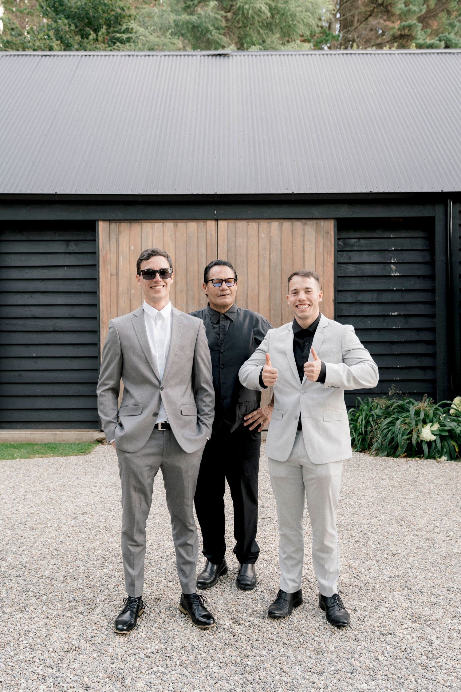 Three groomsmen in formal attire pose together in front of a dark wooden barn.